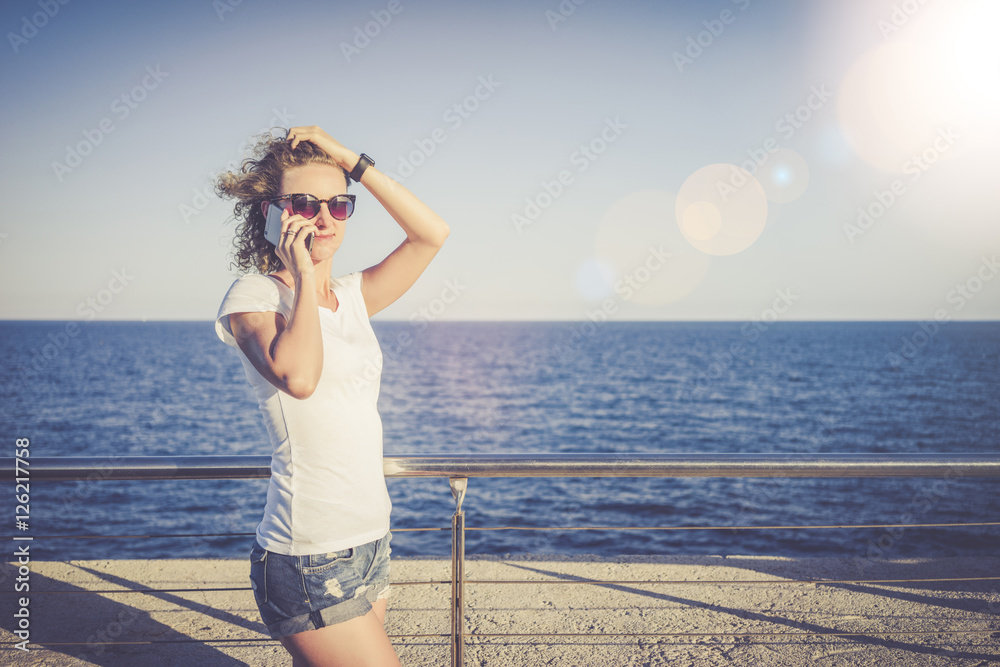 Summer sunny day, a girl dressed in a white T-shirt and denim shorts, standing and talking on cell phone, holding a hand waving hair. In the background the sea. A young woman uses the gadget. Summer sunny day, a girl dressed in a white T-shirt and denim shorts, standing and talking on cell phone, holding a hand waving hair. In the background the sea. A young woman uses the gadget.