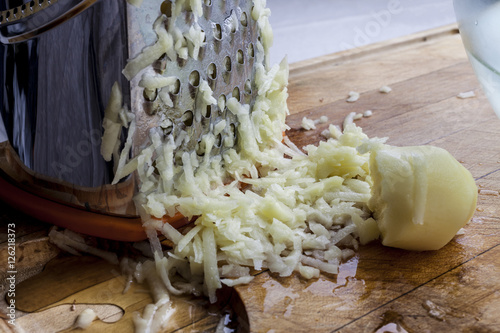 Grated potato with metal standing grater on wooden cutting board from side closeup