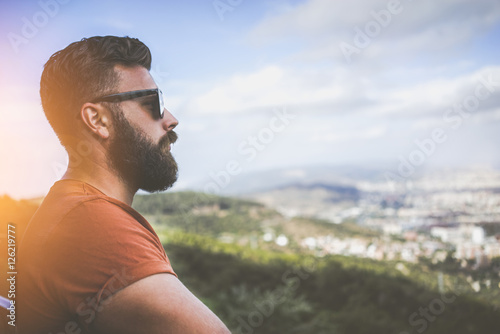 Summer sunny day. Side view of a young bearded man in sunglasses and a brown T-shirt standing on the highest point, looking at the landscape from a bird's flight. Tourist enjoying the view.