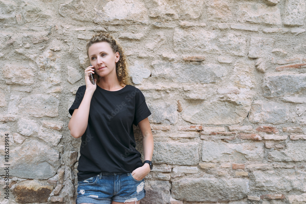 Front view of a young woman dressed in a black T-shirt and denim shorts, standing and talking on the phone. In the background is bright stone wall. Girl uses the gadget. Front view of a young woman dressed in a black T-shirt and denim shorts, standing and talking on the phone. In the background is bright stone wall. Girl uses the gadget.