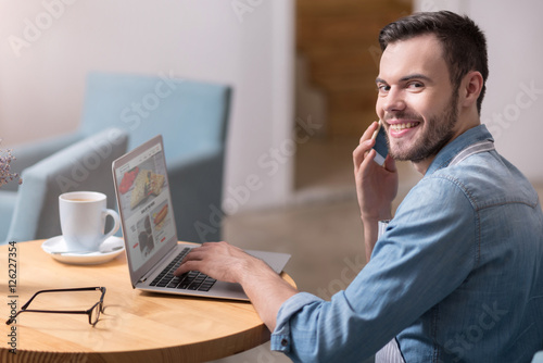 Amused smiling man using laptop and talking on cellphone.