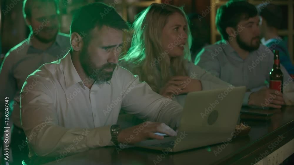 Concentrated middle-aged bearded male worker using a laptop at the bar counter as a young blonde accidently disturbing him and apologizing Concentrated middle-aged bearded male worker using a laptop at the bar counter as a young blonde accidently disturbing him and apologizing