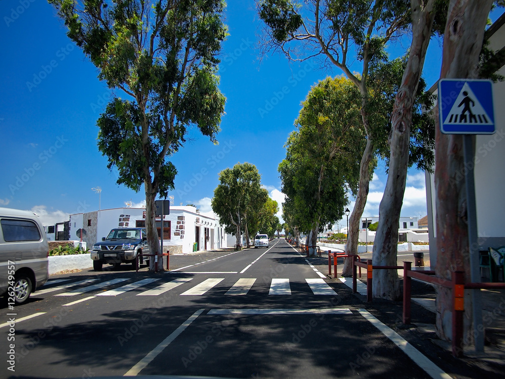 Asphalt road through the southern village with whitewashed houses. White clouds on a blue sky. Lanzarote, Canary Islands, Spain