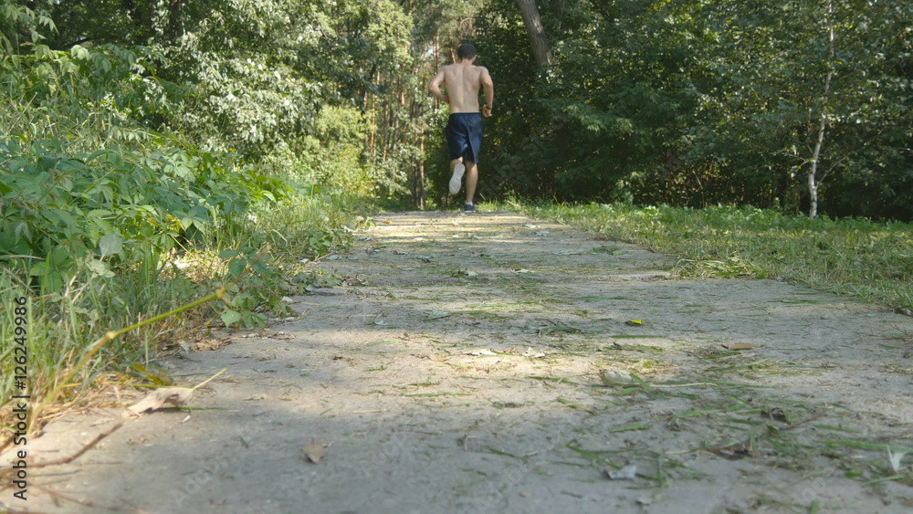 Young muscular athlete running at the forest path. Active strong man training outdoors. Fit handsome athletic male sportsmen working out. Guy training and exercising. Healthy lifestyle. Rear back view