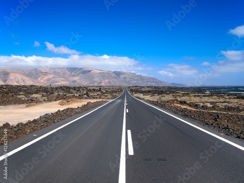 Asphalt road disappearing over the horizon through volcano mountain hillsides. White clouds on a blue sky. Lanzarote, Canary Islands, Spain