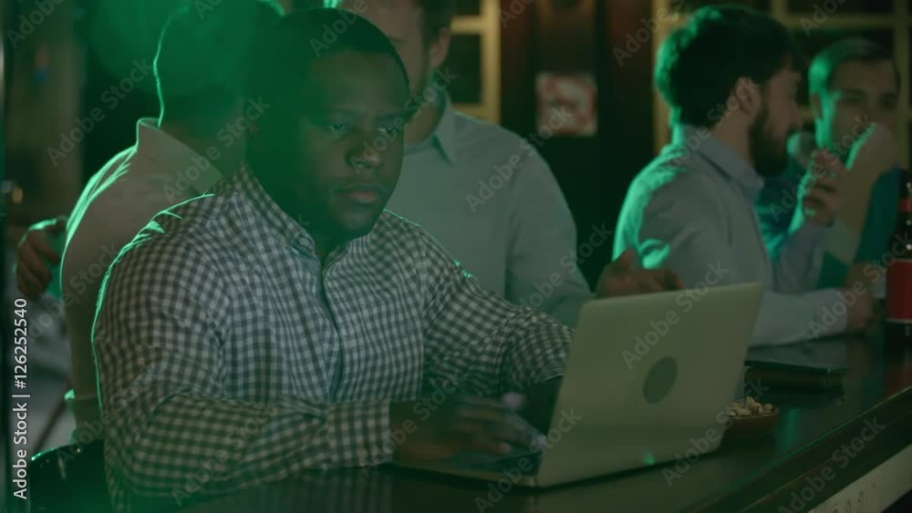 Good-looking concentrated African-American office worker sitting at the bar counter, using a laptop, relaxed drinking bearded coworkers standing next to him Good-looking concentrated African-American office worker sitting at the bar counter, using a laptop, relaxed drinking bearded coworkers standing next to him
