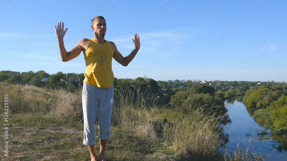 Guy practicing yoga moves and positions outdoors. Young man standing at yoga pose at nature. Athlete doing strength exercise at the hill. Landscape at background. Healthy active lifestyle. Close up Guy practicing yoga moves and positions outdoors. Young man standing at yoga pose at nature. Athlete doing strength exercise at the hill. Landscape at background. Healthy active lifestyle. Close up