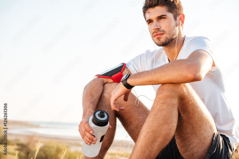 Φωτογραφία Handsome young sportsman sitting and holding bottle of water outdoors