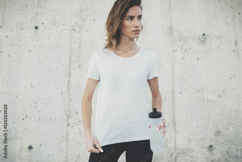 Young fitness girl wearing blank white t-shirt with empty area for design or content, mockup of template white t-shirt, female jogger resting after active workout outside, concrete wall in background Young fitness girl wearing blank white t-shirt with empty area for design or content, mockup of template white t-shirt, female jogger resting after active workout outside, concrete wall in background