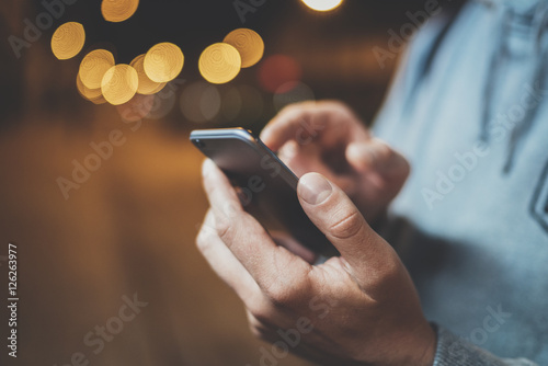 Closeup image of male hands using modern smartphone at night, bokeh light in blurred background, young hipster guy typing sms message on touch screen of cellphone, social networking concept
