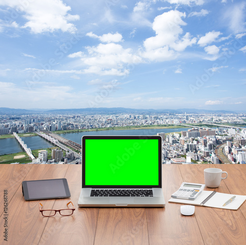 Blank green screen laptop computer on wooden table with cityscape background
