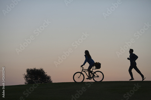 Silhouette of a couple walking through a park in San Diego at dusk. The woman is riding a bike and the man is walking. 