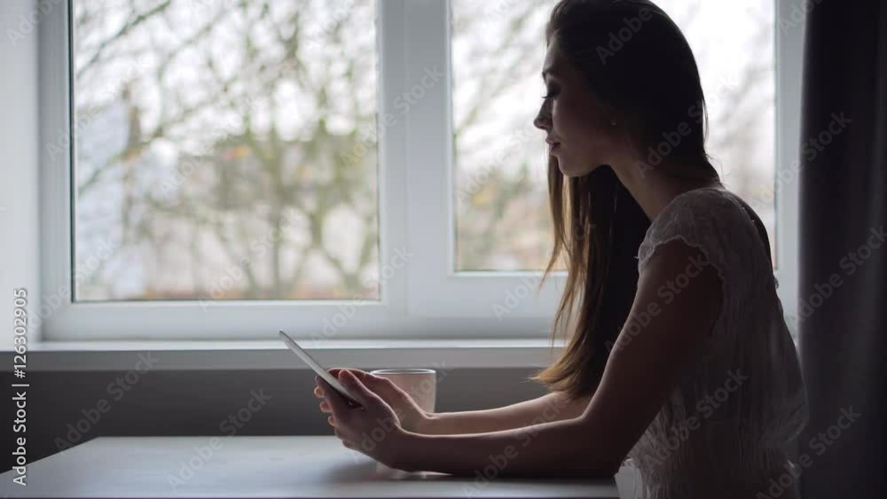 A attractive young brunette girl works with tablet pc in kitchen near window A attractive young brunette girl works with tablet pc in kitchen near window