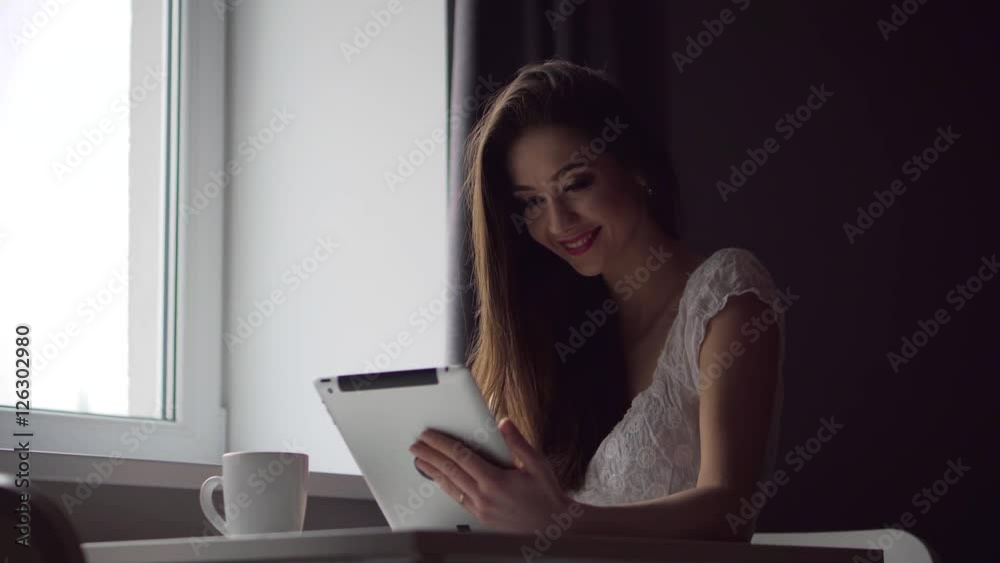 A attractive young brunette girl works with tablet pc in kitchen near window A attractive young brunette girl works with tablet pc in kitchen near window