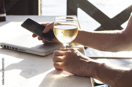 Cropped image of man's hand with telephone and glass of white wine near the sea.