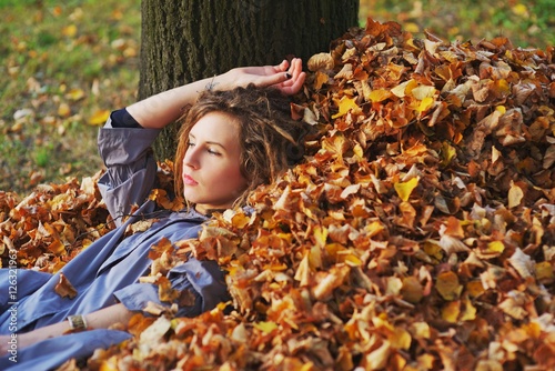 Cute teen girl in a light purple cloak with sad eyes lies on a pile of fallen autumn leaves in the forest  leaning his head against the tree  close up.