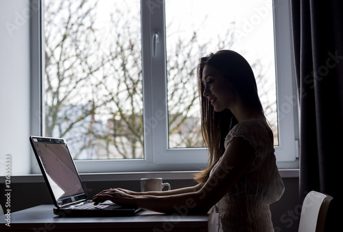 A attractive young brunette girl works with laptop in kitchen near window © proimagecontent