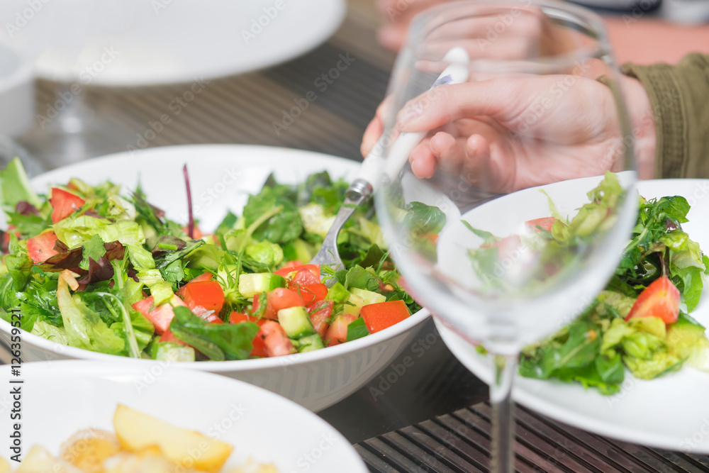 Empty wine glass, a human hand with a spoon and vegetable salad with tomatoes and greens on a white plate
