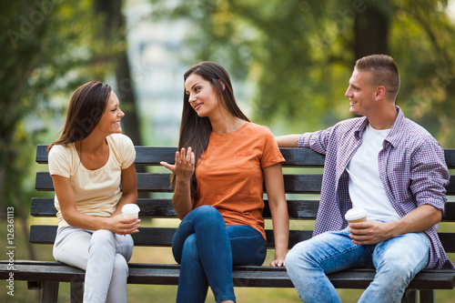 Three friends are sitting on bench in park and talking.