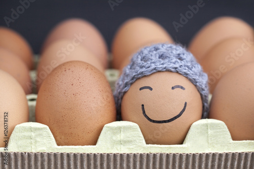 Smiling egg wearing a knitted hat souronded by blank brown eggs.