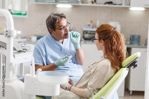 Fototapeta Naklejka Na Ścianę i Meble -  A professional dentist, equipped with a bright smile, converses with his red-haired female patient, carefully explaining the upcoming treatment, and ensuring her comfort throughout the process.