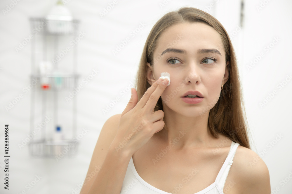 Young beautiful woman applying cream to her face and looking worried at the mirror at home bathroom. Beauty, skin care concept, lifestyle Young beautiful woman applying cream to her face and looking worried at the mirror at home bathroom. Beauty, skin care concept, lifestyle