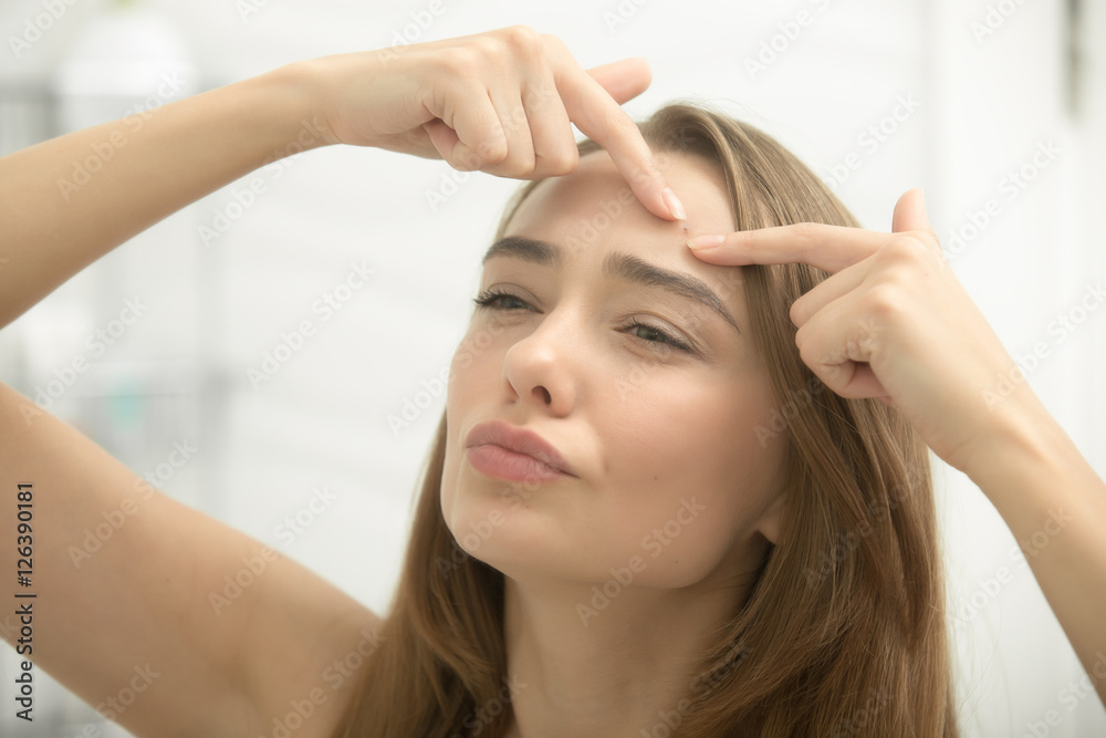 Young woman troubled checking acnes on her forehead, looking at the bathroom mirror. Beauty, skin care concept, lifestyle, close up Young woman troubled checking acnes on her forehead, looking at the bathroom mirror. Beauty, skin care concept, lifestyle, close up