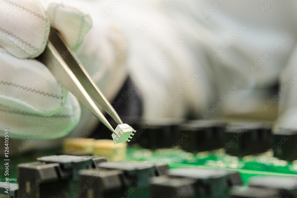 Fototapeta premium Female hand in ESD gloves holding tweezers and assembling white microchip on printed circuit board
