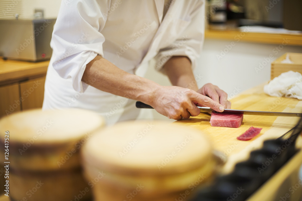 A chef working in a small commercial kitchen, an itamae or master chef slicing fish with a large knife for making sushi A chef working in a small commercial kitchen, an itamae or master chef slicing fish with a large knife for making sushi