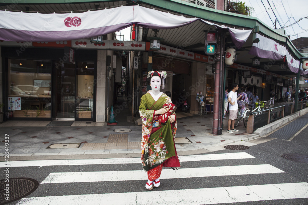 A woman dressed in the traditional geisha style, wearing a kimono and obi, with an elaborate hairstyle and floral hair clips, with white face makeup with bright red lips and dark eyes crossing a street. A woman dressed in the traditional geisha style, wearing a kimono and obi, with an elaborate hairstyle and floral hair clips, with white face makeup with bright red lips and dark eyes crossing a street.