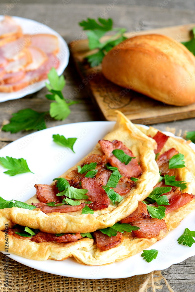 Homemade bacon omelet. Delicious omelet stuffed with bacon and parsley on a plate, bacon slices on a plate, bread roll, fork, knife, cutting board, fresh parsley sprigs on old wooden table. Closeup Homemade bacon omelet. Delicious omelet stuffed with bacon and parsley on a plate, bacon slices on a plate, bread roll, fork, knife, cutting board, fresh parsley sprigs on old wooden table. Closeup