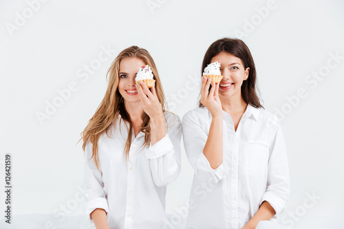Two cheerful girlfriends holding sweet muffins while sitting in bed