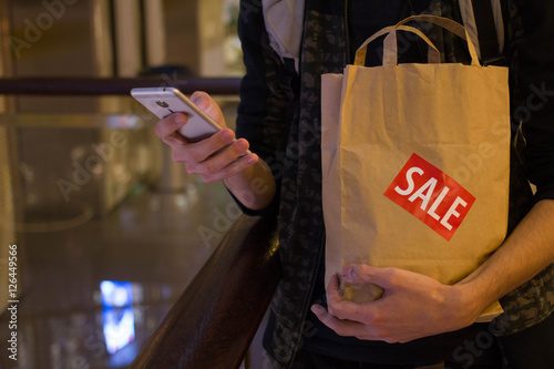 Man hold brown paper bag with SALE sticker on it in one hand and use smartphone with another in mall photo