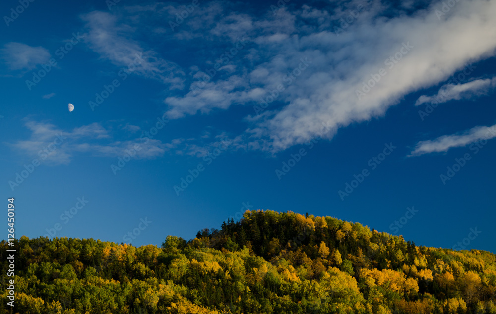 Fall landscape with a mountain top, clouds and the moon in daylight