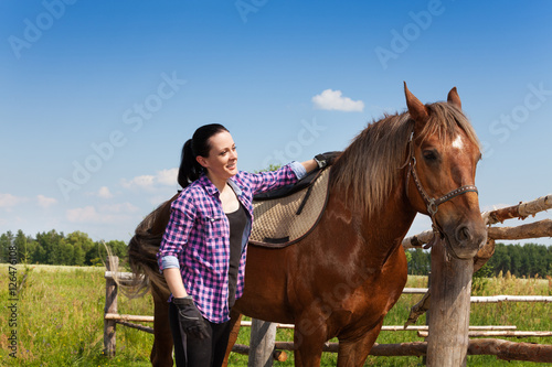 Happy young woman saddling a chestnut brown horse