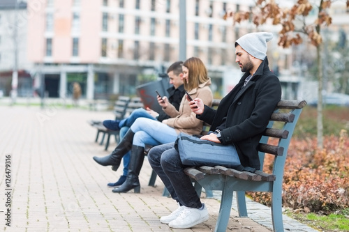 Group of friends sitting on a bench in park separately looking at their phones loosing communication.