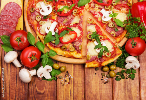 Pizza and slice with salami, mushrooms, vegetables, against dark wood.