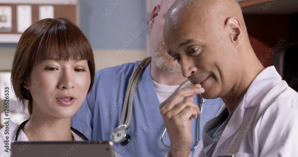 Male nurse and two doctors consult with each other while looking at information on tablet computer in hospital exam room. Close up with nurse center. Male nurse and two doctors consult with each other while looking at information on tablet computer in hospital exam room. Close up with nurse center.