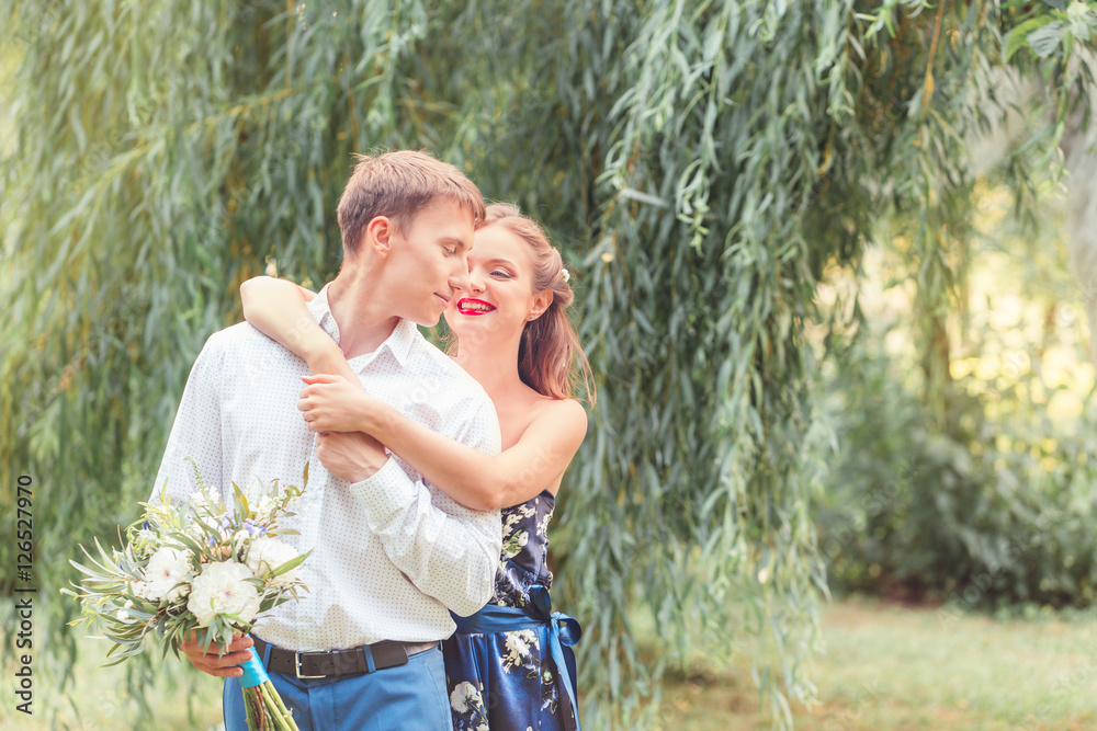 Naklejka premium Happy young couple, she hugs him from behind. Holding a bouquet.