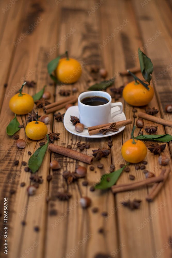 Small white cup of coffee, cinnamon sticks, cocoa beans, star anise, hazelnuts and mandarins on wooden background Small white cup of coffee, cinnamon sticks, cocoa beans, star anise, hazelnuts and mandarins on wooden background