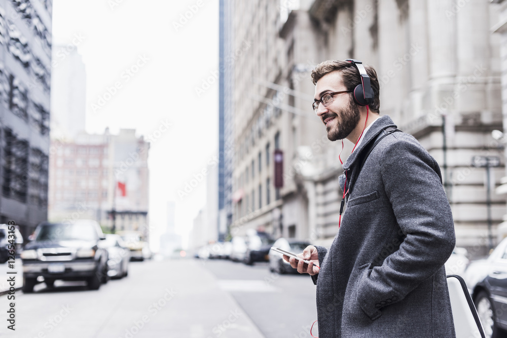 USA, New York City, smiling businessman with cell phone and headphones on the go USA, New York City, smiling businessman with cell phone and headphones on the go