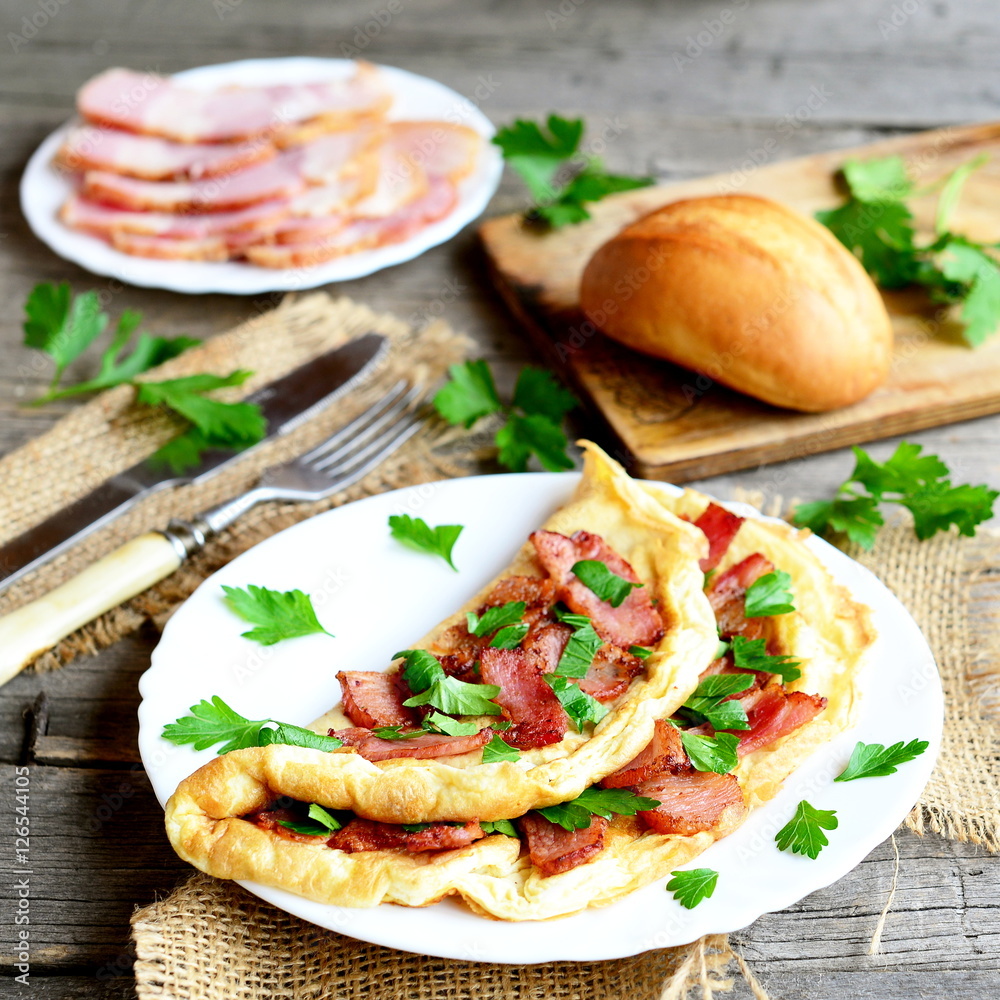 Delicious bacon omelette. Omelette stuffed with bacon and parsley on a plate, bacon slices on a plate, bread roll, fork, knife, chopping board, fresh parsley on old wooden background. Homemade cuisine Delicious bacon omelette. Omelette stuffed with bacon and parsley on a plate, bacon slices on a plate, bread roll, fork, knife, chopping board, fresh parsley on old wooden background. Homemade cuisine