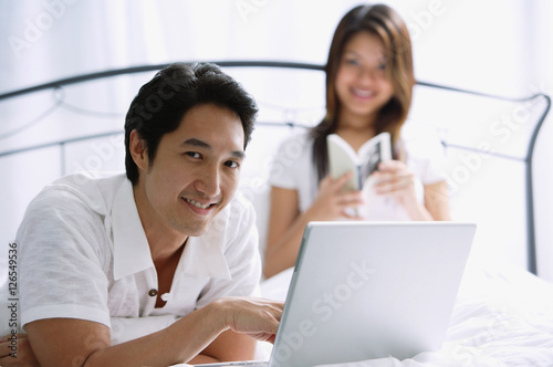 Couple in bedroom, man using laptop, looking at camera © Alexander