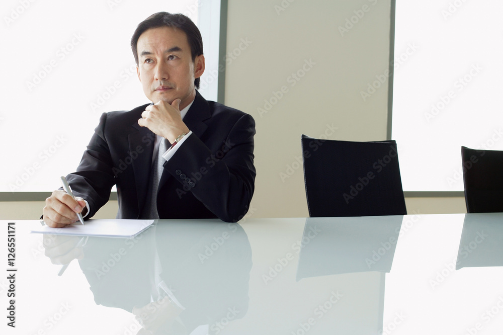 Businessman sitting at table, with pen and paper, hand on chin Businessman sitting at table, with pen and paper, hand on chin