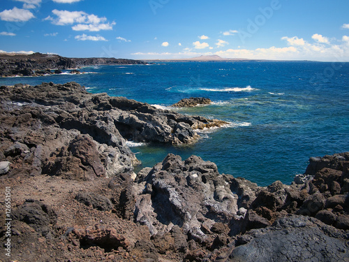 Ocean waves breaking on the rocky coast of hardened lava with caverns and cavities. Deep blue sky with white clouds and mountains and volcanoes on the horizon. Lanzarote, Canary Islands, Spain