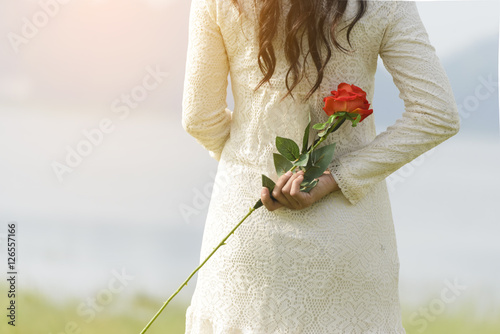 Beautiful women wearing white dress standing behind holding a red rose.A woman looking at the lake in front of her.