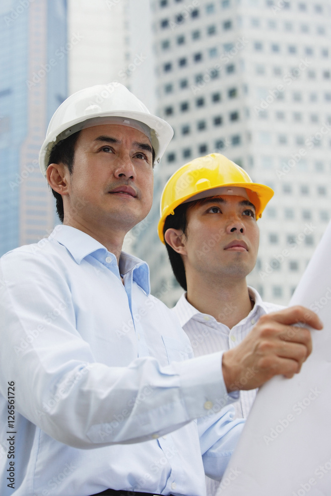 Men in hardhats, holding blueprints, buildings in the background Men in hardhats, holding blueprints, buildings in the background