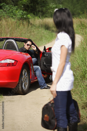 Woman standing on side of road, carrying bag, looking at man in red sports car © Alexander