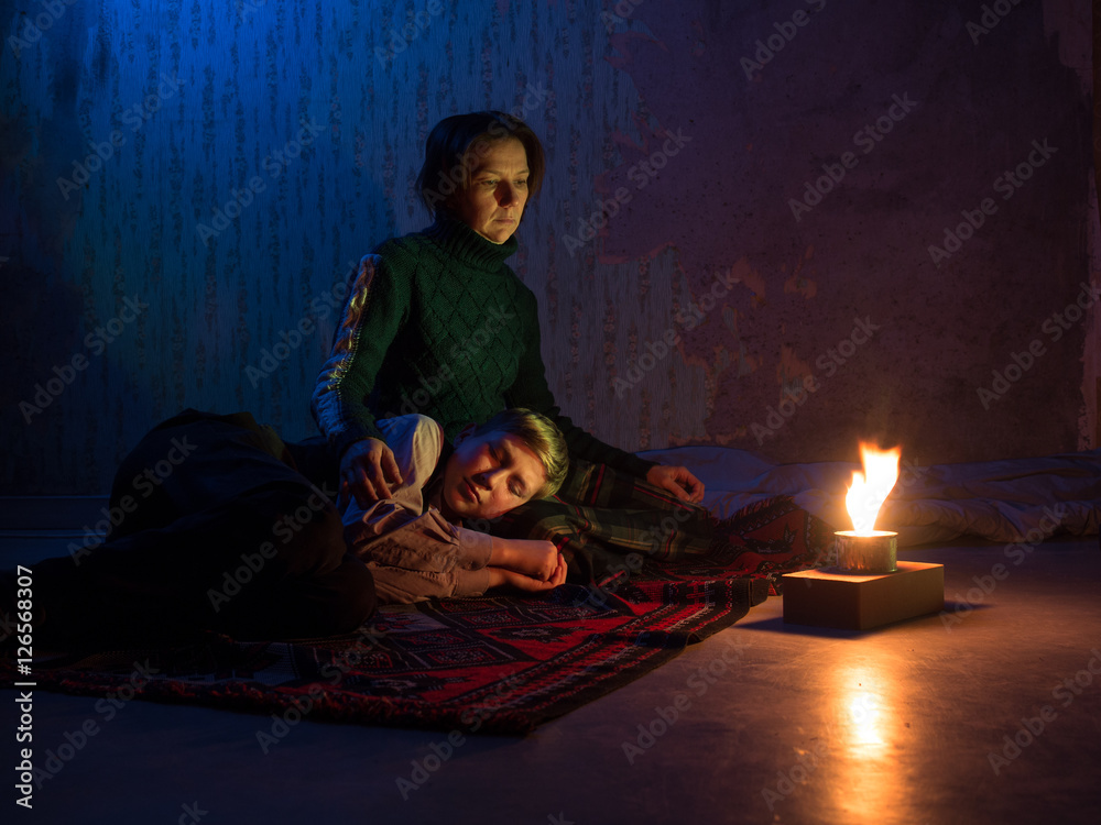 Fototapeta premium Adult woman sitting on the floor and sadly looks and the young boy sleeping on her lap against the backdrop of concrete walls with torn wallpaper and a burning candle