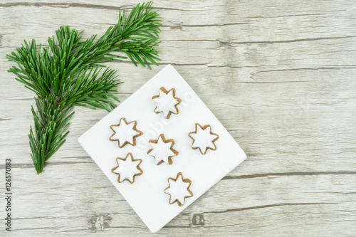 Kleiner quadratischer weißer Teller mit Weihnachtsplätzchen, Zimtsternen, einem Tannenzweig, Untergrund aus Holz, Textfreiraum